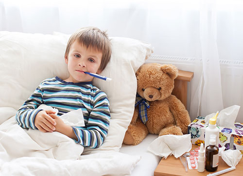 little boy laying in bed with his teddy bear and with a thermometer in his mouth, staying home from school because he is sick
